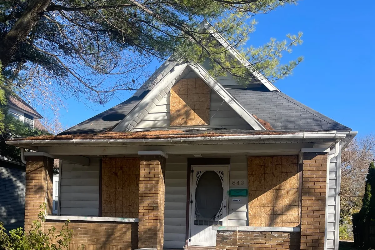 Before the renovation. A small bungalow with boarded up windows, a worn brick porch, and an overgrown front yard.